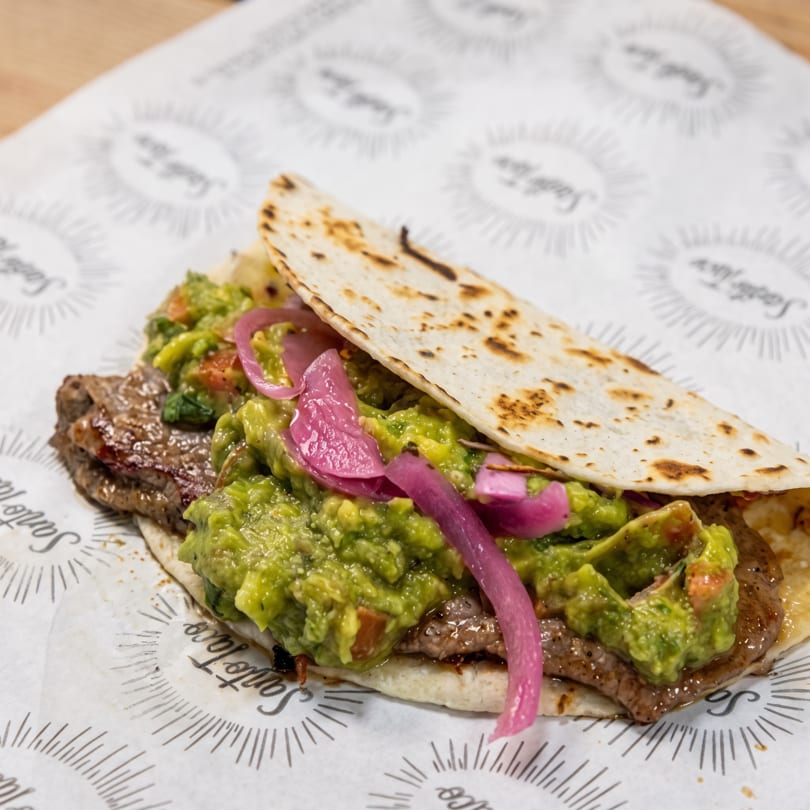 A close-up of a Santo Taco with grilled meat, guacamole, and pickled red onions on a tortilla.
