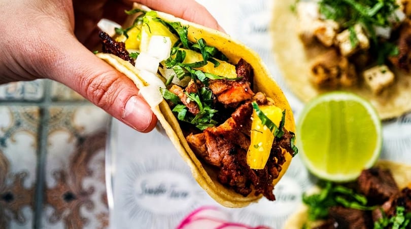 Hand holding a taco filled with meat, pineapple, cilantro, and onions, with a lime in the background.