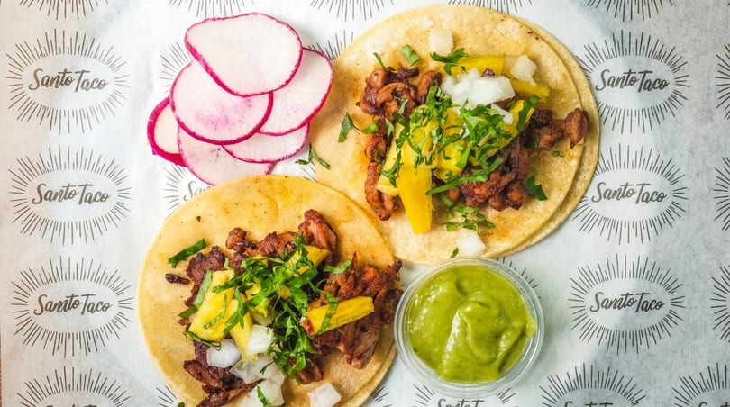 Two Santo Taco al pastor tacos with pineapple, onions, cilantro, radishes, and guacamole.