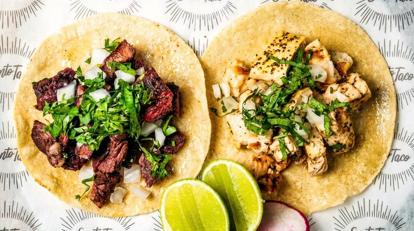 Two tacos, one with steak and one with chicken, topped with onions and cilantro, with lime wedges.