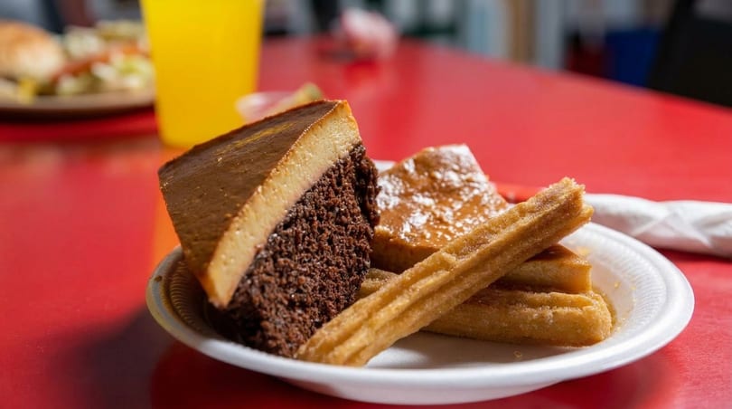 A plate of chocolate flan cake and churros on a red table with a drink in the background.