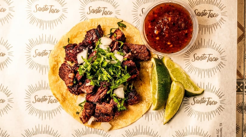 A Santo Taco carne asada taco with onions and cilantro, salsa, and lime wedges.
