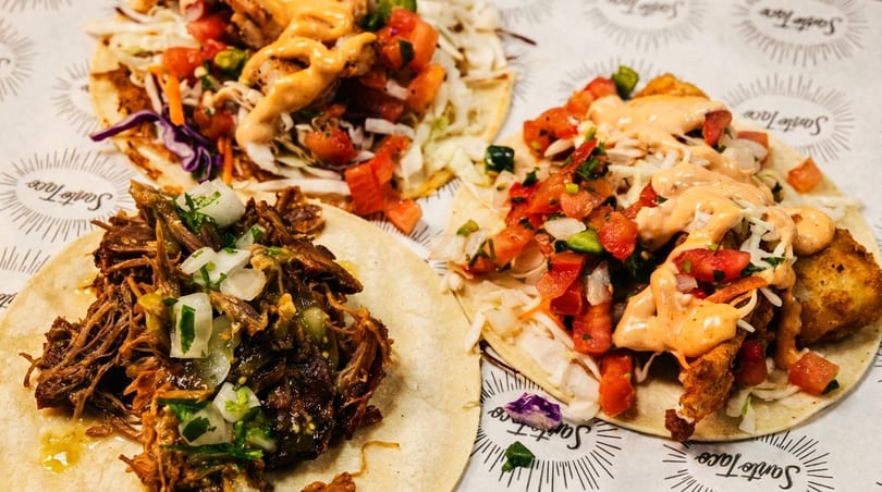 Three Santo Taco tacos, one with shredded beef, two with fried fish, salsa, and sauce.