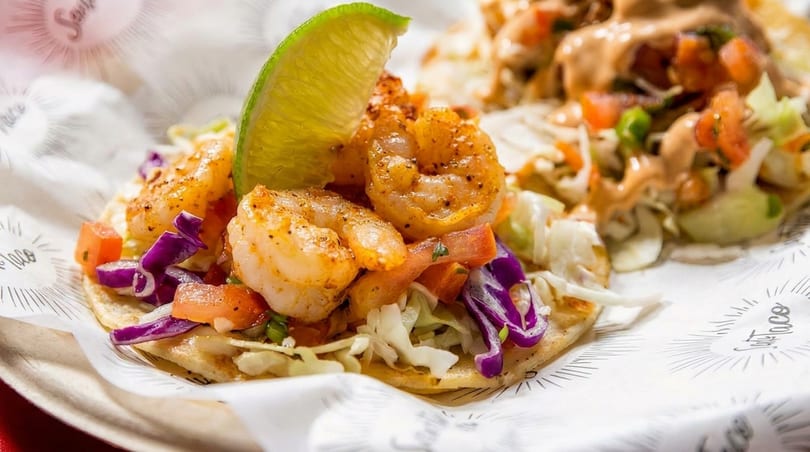 Two shrimp tacos with cabbage, pico de gallo, and a lime wedge on branded paper.