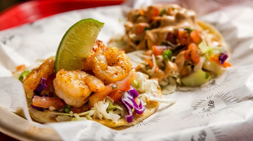 Two shrimp tacos with cabbage, tomatoes, and lime wedges on branded paper.