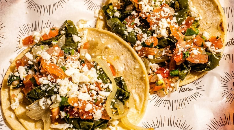 Two vibrant tacos filled with green vegetables, tomatoes, onions, and crumbled white cheese on patterned paper.